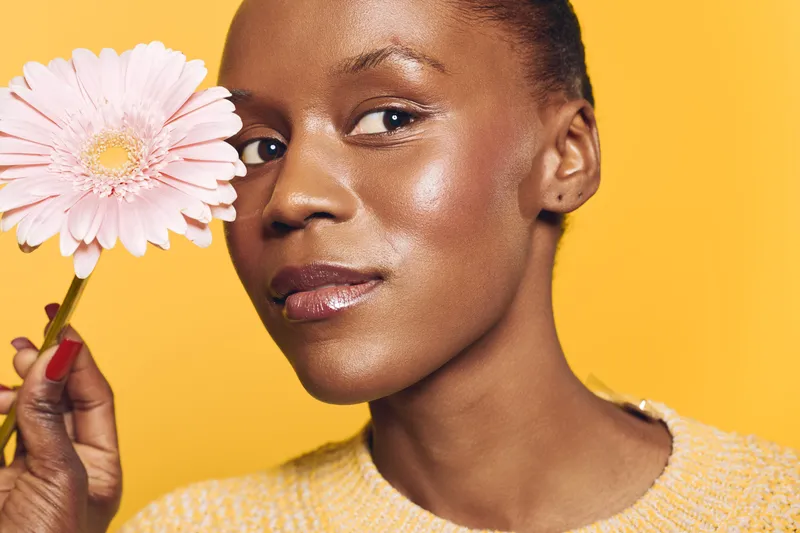 Close-up of a confident African woman with dark skin holding a pink flower near her face against a yellow background. Concept of natural beauty, skincare, and wellness.