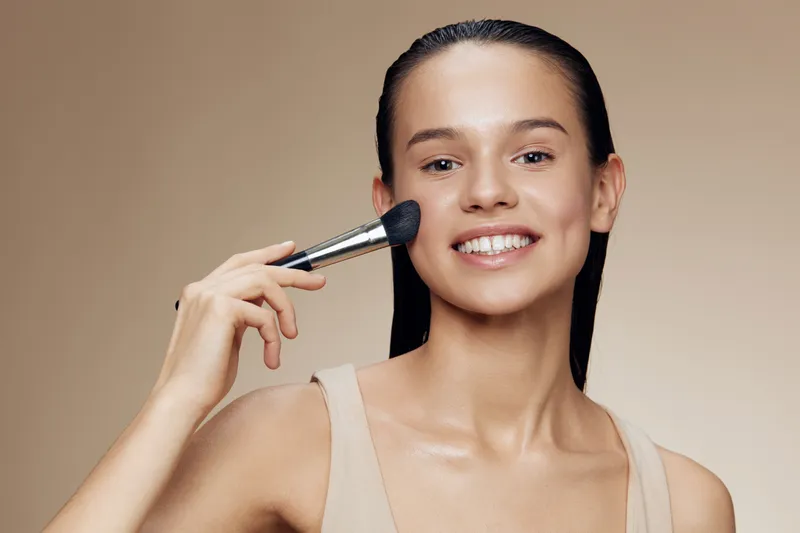 Smiling young woman applying makeup with a brush on a neutral background, showcasing beauty and confidence, perfect for themes of skincare and self care