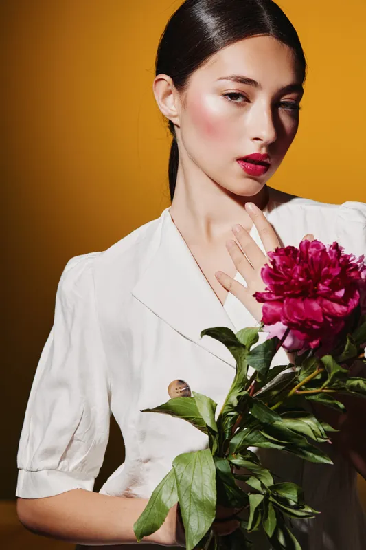 Young woman with glossy dark hair poses in white blouse holding vibrant peony flower. Fashion portrait with creative makeup and elegant styling on orange background. Beauty concept.