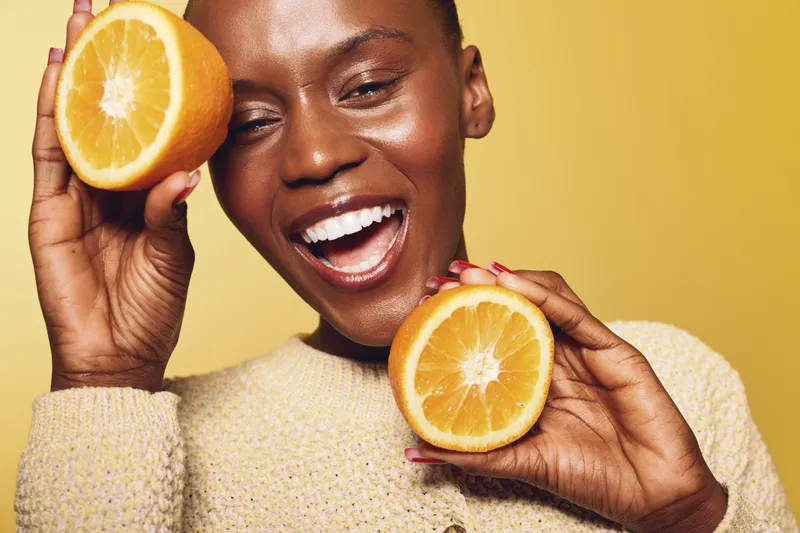 Happy African woman with dark skin holds two orange halves near her face smiling with closed eyes on yellow background. Concept of healthy lifestyle, vitamin, and natural beauty care.