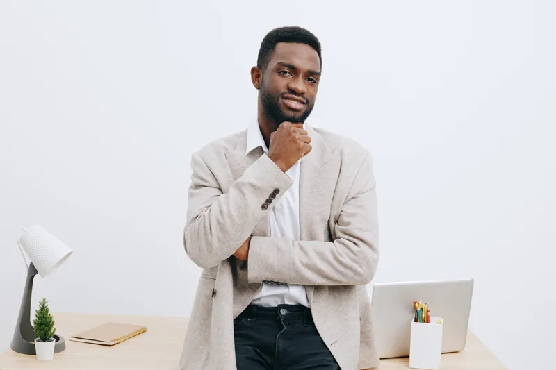 Confident young black man dressed in casual beige blazer and white shirt standing in modern office environment holding chin pose looking at camera isolated on plain white background studio portrait pe