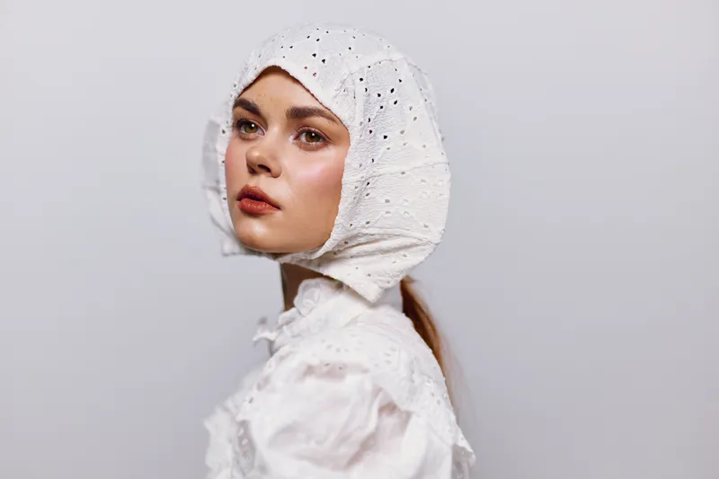 Young woman in a white bonnet and blouse, showcasing serene emotions against a light gray background, emphasizing vintage beauty and delicate style