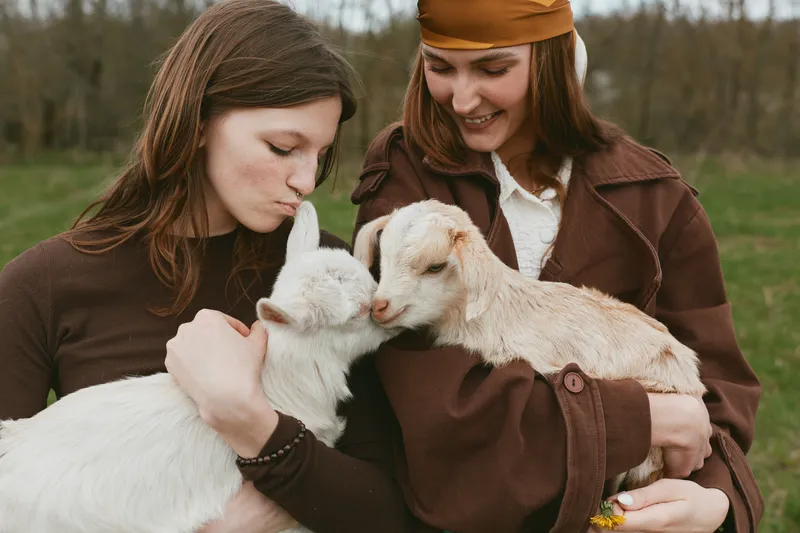 Goat, kid, lifestyle, outdoor scene with two women embracing gentle animals in a sunlit field, candid interaction, warm textures, casual outfits, rustic backdrop, wholesome companionship, nature