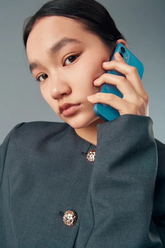Portrait of young woman with smartphone holding phone to ear for communication and call. Female user in formal jacket with serious expression using mobile device against plain background.