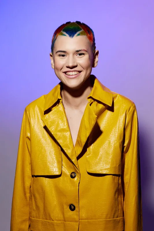 young woman with colorful short hair wearing a yellow jacket smiling confidently against a purple gradient background, modern fashion portrait, vibrant style