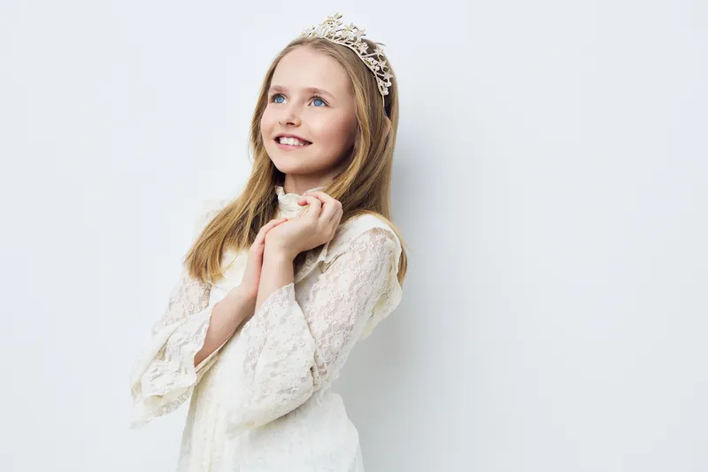 Sweet young girl with a tiara in a white dress smiling happily against a soft white background, conveying joy and innocence in a serene atmosphere