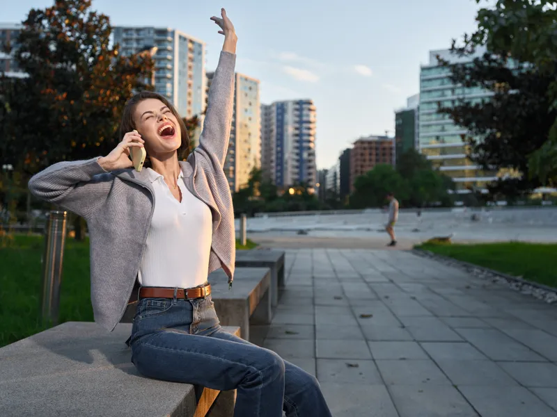 A cheerful woman sits on a park bench in an urban plaza, talking on a phone with a bright smile, enjoying a sunny day and a vibrant city atmosphere surrounding her.