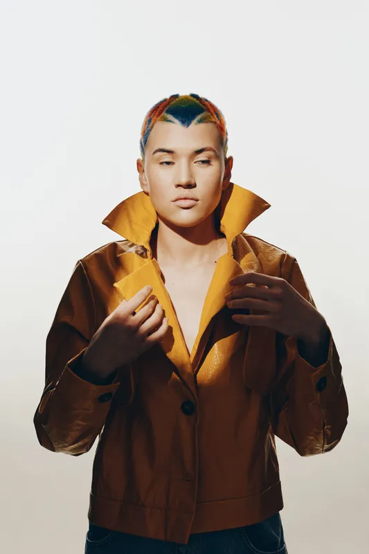 woman with colorful braided hair wearing brown jacket, standing confidently against plain light background, portrait with unique hairstyle and fashion style.