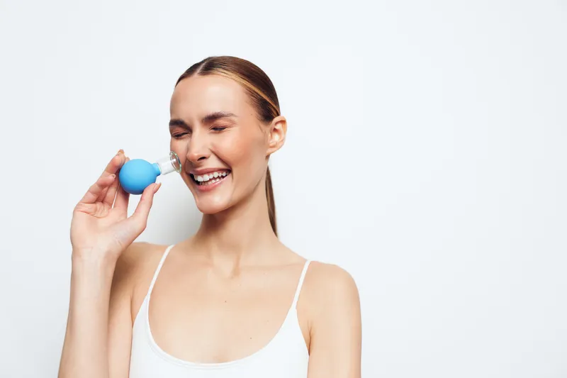 Young woman smiling with eyes closed using a facial cleansing device on white background, fresh skin care routine, natural light, clean and simple composition.