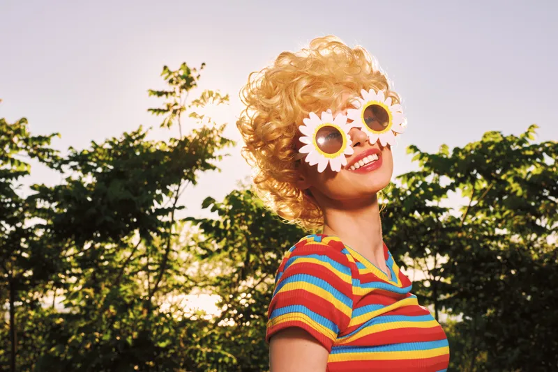 Smiling woman with curly hair wearing a colorful striped top and flower sunglasses outdoors under warm sunshine, capturing a vibrant lifestyle with retro flair and carefree mood.