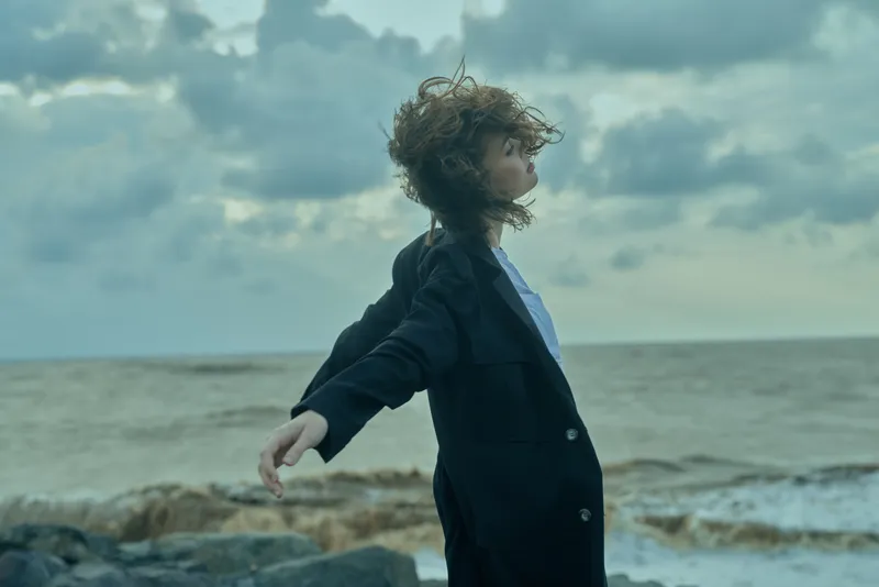Business, wind, sea, jacket scene with a figure standing on a rocky shore, coat fluttering, open posture and expansive horizon, empowering mood.