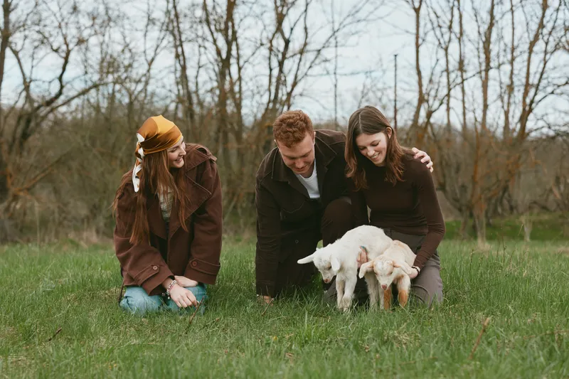 Three friends outdoors with a playful goat kid, kneeling on green grass, sharing a warm candid moment in a natural field, enjoying animal companionship and relaxed lifestyle.