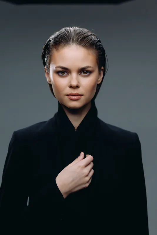 serious woman with slicked hair wearing black jacket posing on gray background