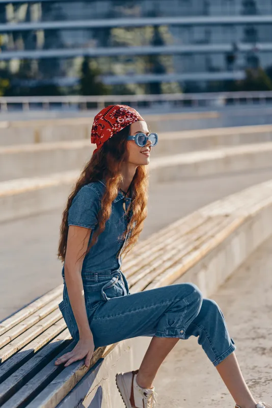 young woman sitting outdoors wearing sunglasses and a red bandana, dressed in blue denim and casual shoes enjoying sunny weather on wooden benches