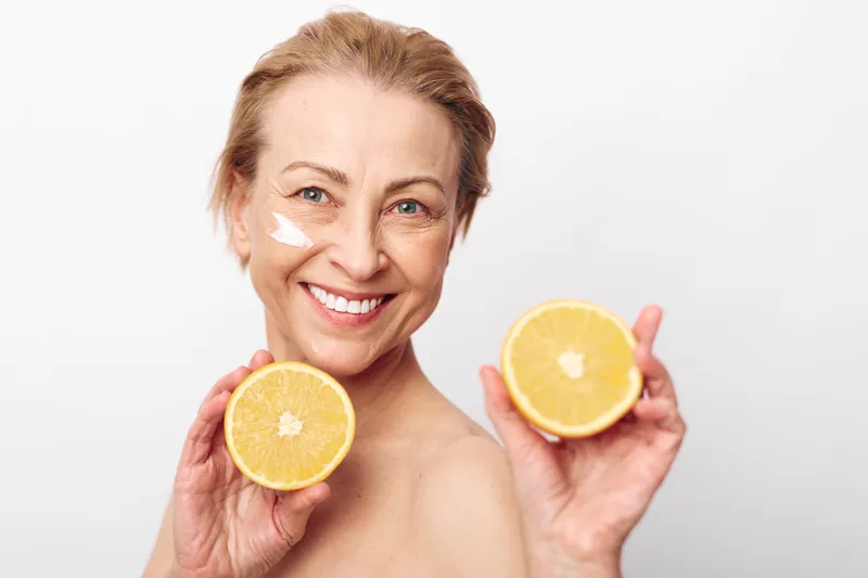 Smiling mature woman with natural skin holds two halves of fresh orange close to face with cream applied on cheek. Concept of healthy lifestyle, skincare, citrus fruits, wellness and self-care.
