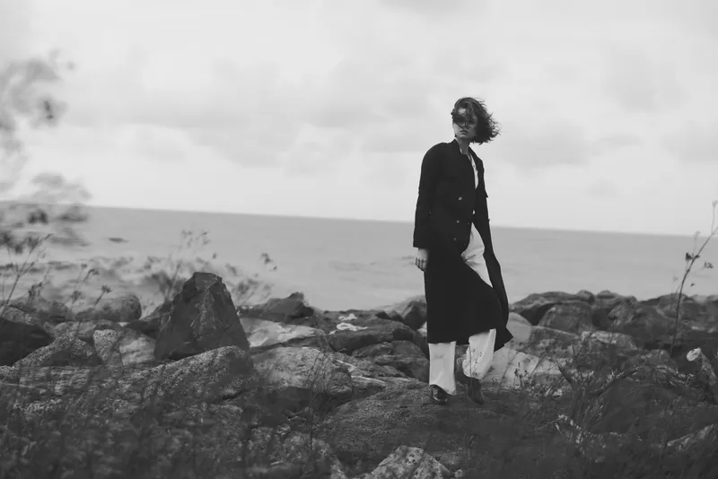 Monochrome scene of a woman standing on rugged rocks by the sea, windblown coat, solitary mood, dramatic coastline and open sky
