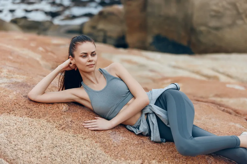 Athletic young woman in stylish activewear relaxing on rocky beach, portraying confidence and a healthy lifestyle surrounded by nature
