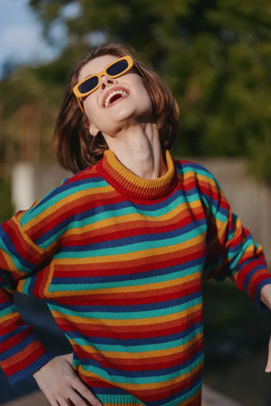 Young woman wearing a colorful striped sweater and yellow sunglasses laughing outdoors, short hair, casual outfit, leaning back in sunlit park for lifestyle fashion and travel mood.