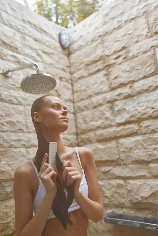 woman showering outdoors with wet hair and white swimwear under natural light at stone wall background, summer wellness moment