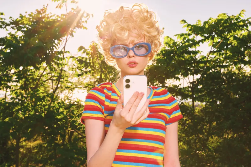 Cheerful woman outdoors, wearing a colorful striped shirt and retro sunglasses, taking a selfie in bright sunlight, relaxed pose, natural background, warm tones, vintage mood, lively moment