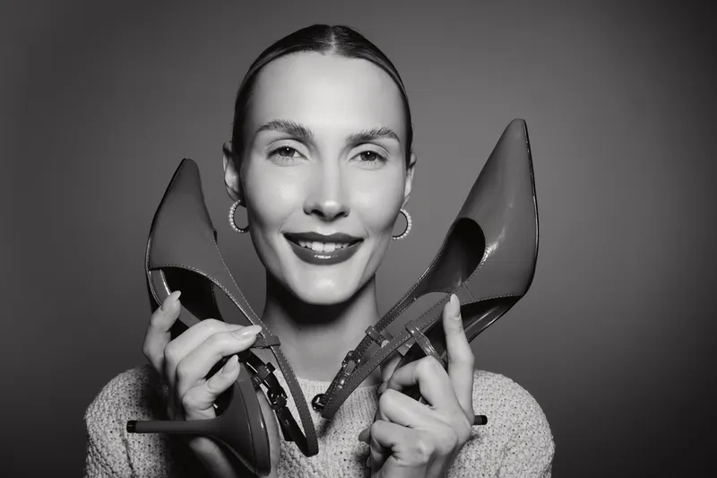 Smiling young Caucasian woman with fair skin holding high heel shoes near face, wearing earrings and sweater. Portrait in black and white studio lighting, fashion and style concept.