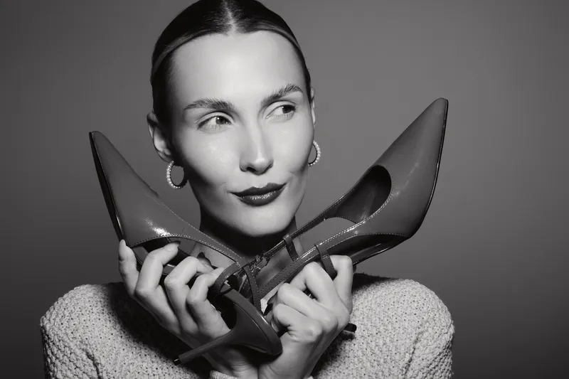Young Caucasian woman with pale skin and slicked back hair holds red high heels near her face, posing in studio with neutral background. Concept of fashion and style.