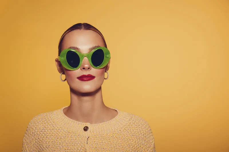 Stylish woman with slicked back hair wears round green sunglasses and yellow knitted sweater against yellow background. Concept of fashion, summer style, and modern accessories.
