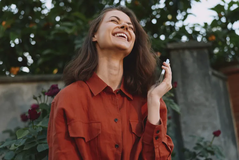 Woman lifestyle, outdoor moment, laughing with head tilted back, red shirt, phone in hand, sunlit garden backdrop, candid relaxed vibe, natural colors
