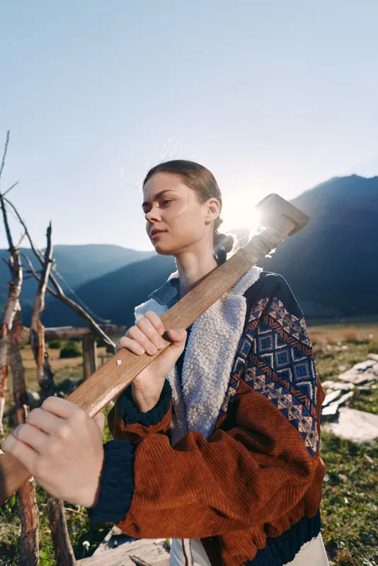 Woman with axe and wood in mountains outdoors, rural scene of chopping and nature. Female lumberjack portrait shows focused expression, sunlight, jacket and hands on handle.