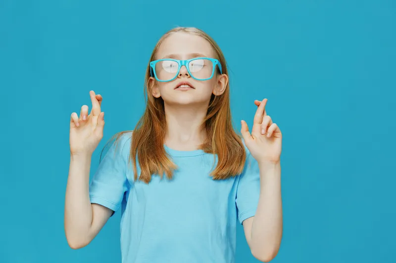 Young girl with glasses crossing fingers, displaying hope and optimism against a bright blue background, representing positivity and childhood dreams
