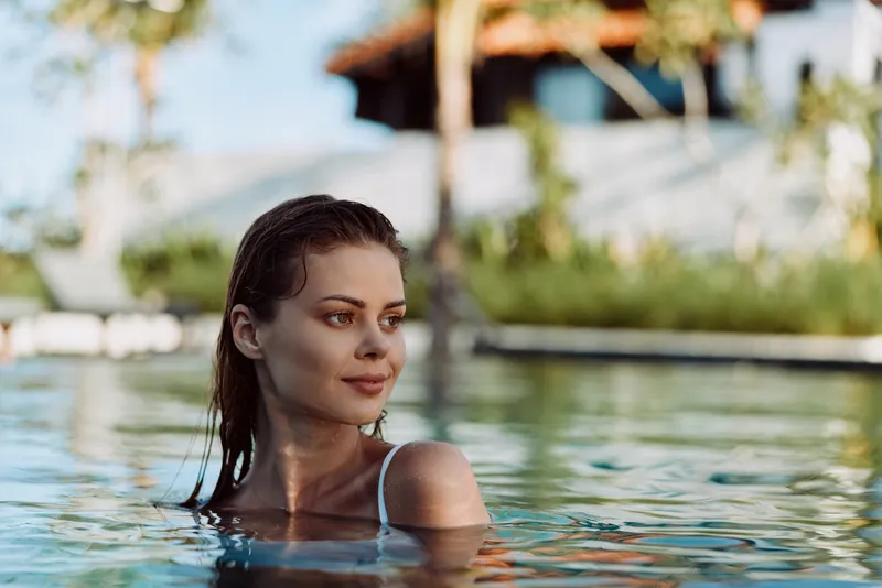 A calm young woman with wet hair relaxes in a sunlit pool, looking to the side as water glistens around her shoulders, capturing a candid moment of leisure