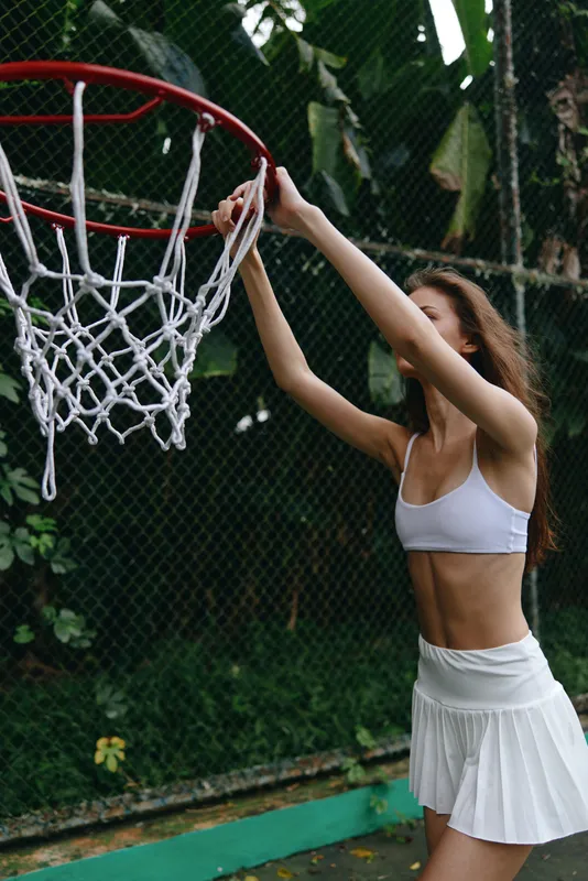 Athletic young woman in a white sports outfit shooting basketball, showcasing energy and determination in a vibrant green outdoor court