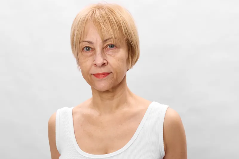 Elderly woman portrait, short hair blond, wearing white top, studio background, calm neutral expression, mature face closeup, soft light, copyspace, for health care