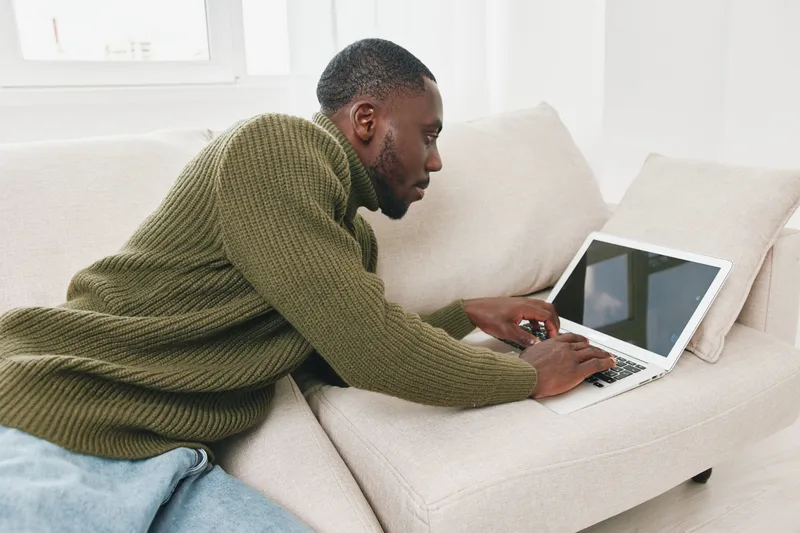Young black man working on a laptop at home, wearing a knitted green sweater, relaxed and focused, ideal for remote work and productivity concepts