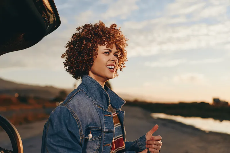 Happy woman lifestyle portrait outdoors, curly hair, wearing denim jacket, smiling and gesturing on a road at sunset, cinematic warm light, relaxed travel mood, lifestyle photography