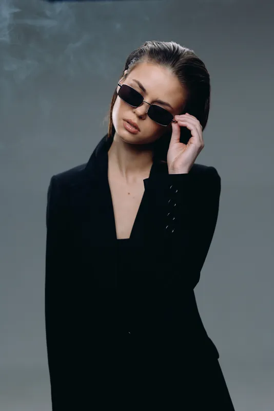 Stylish woman wearing sunglasses and black blazer posing with confidence in a minimalist studio setting.