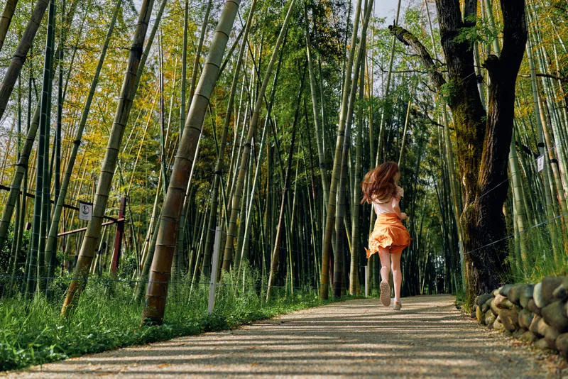 Girl in orange dress running barefoot on a forest path through tall bamboo and trees, sunlight filtering through leaves creating a serene nature scene of motion and summer freedom.