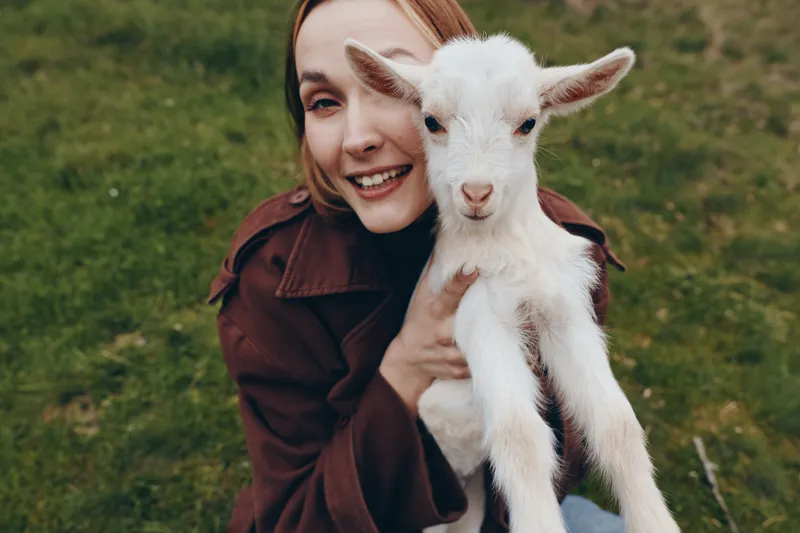A smiling woman outdoors cuddles a white goat kid in a green field, capturing a lifestyle moment of bonding with animals and rural calm amid nature.