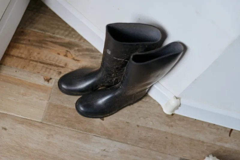 A pair of black boots rests on a wooden floor beside a white wall, creating a minimal indoor scene that highlights everyday footwear left by the door in a cozy home environment.