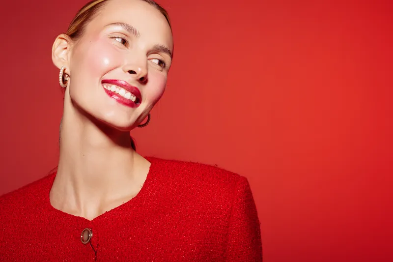 Smiling woman with red lipstick and red outfit looks away on red background. Studio portrait with bright makeup and earrings. Concept of fashion, beauty, and confidence.