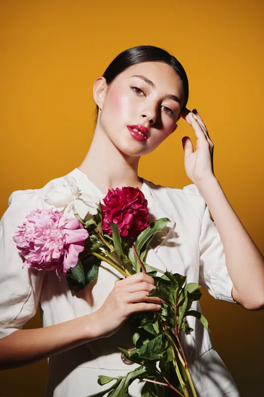 Young woman with glossy dark hair holds pink and red flowers against a warm mustard background. Her flawless skin glows with natural makeup and glossy red lips in fashion style.
