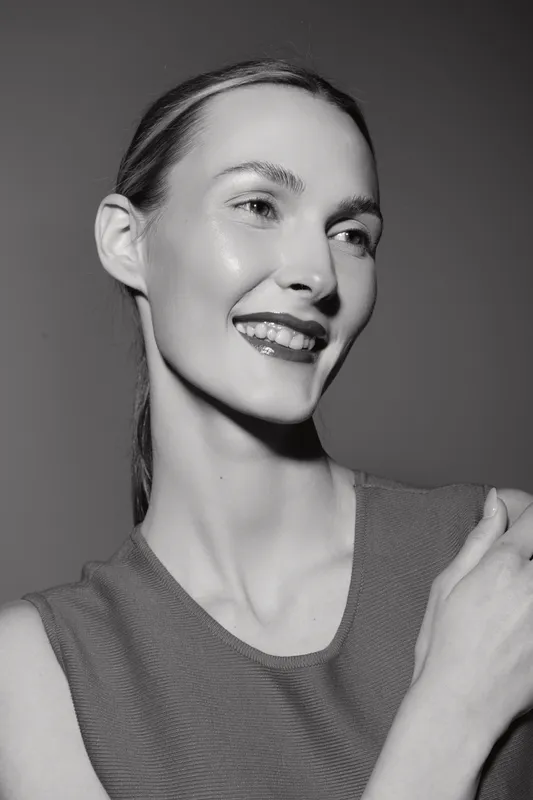 Smiling caucasian woman with light skin and slicked back hair in a sleeveless top looks to side in black and white studio portrait.