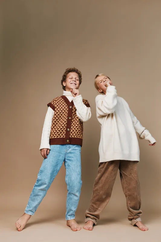 Two playful children pose together in cozy knitwear and relaxed denim, barefoot on a neutral backdrop, showcasing warm fashion and carefree joy in a bright, studio style.