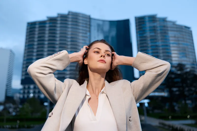 confident woman in beige coat posing in urban business district with modern skyscrapers background at dusk