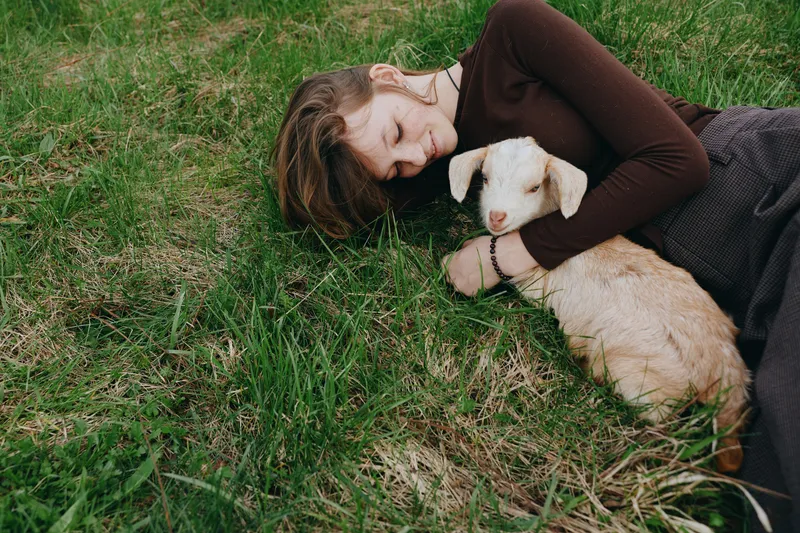 Calm woman lying on grass sharing a tender moment with a goat kid, embracing nature and a relaxed lifestyle. Soft light highlights affection, outdoor serenity, and warm well being.