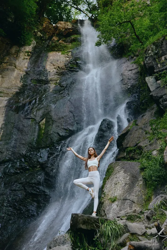 young woman practicing yoga pose near a tall waterfall in lush green forest, peaceful nature, outdoor activity, wellness and balance