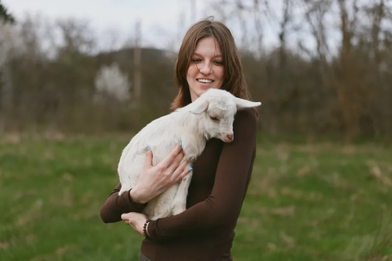 A smiling woman outdoors holds a young goat kid in a grassy field, presenting a warm, gentle lifestyle moment. The scene emphasizes bonding, care, and natural companionship between human and animal.