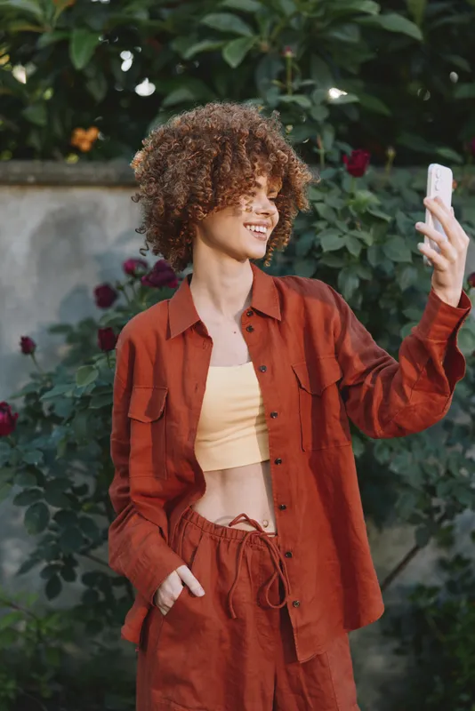 Smiling woman with curly hair taking a selfie outdoors in a rust orange outfit. Lush garden backdrop. Focus on lifestyle fashion and self expression through photography. Soft daylight highlights