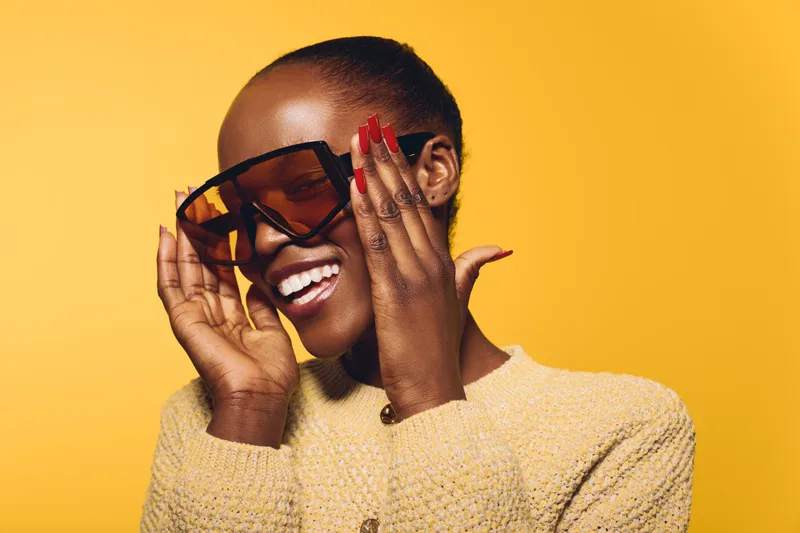 Smiling dark skinned African woman wearing large sunglasses and yellow sweater poses against yellow background. Concept of fashion, style, beauty, and confident young black female model.