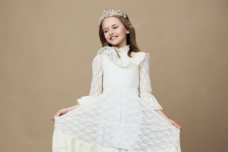 Young girl in a beautiful white dress with lace, smiling joyfully, wearing a crown against a plain beige background, perfect for seasonal celebrations and children s fashion